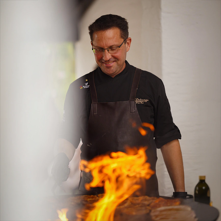 Jürgen Reck steht lächelnd an einer Feuerplatte und blickt auf das Grillgut, aus dem helle Flammen emporschlagen. Er trägt eine Brille, eine schwarze Kochjacke mit Stickerei „Dorfmetzger“ und eine dunkelbraune Lederschürze. Rechts neben der Grillplatte steht eine Flasche Öl, im Hintergrund eine weiß getünchte Wand.