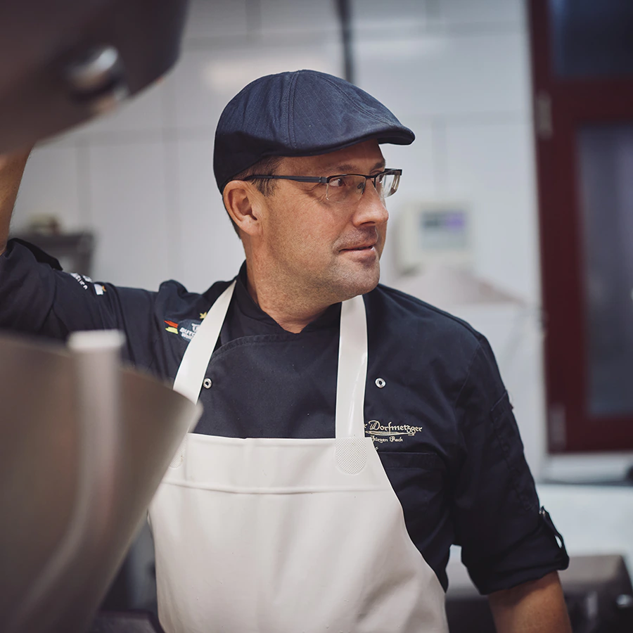 Jürgen Reck, der Dorfmetzger, in seiner Metzgerei-Küche. Er trägt eine dunkelblaue Schiebermütze, eine Brille, eine schwarze Kochjacke mit goldener Stickerei „Dorfmetzger Jürgen Reck“ und eine weiße Schürze. Mit einem Arm stützt er sich auf eine große Edelstahlmaschine im Vordergrund und blickt mit leichtem Lächeln zur Seite. Im Hintergrund sind weiß geflieste Wände und ein rot gerahmtes Fenster zu sehen.